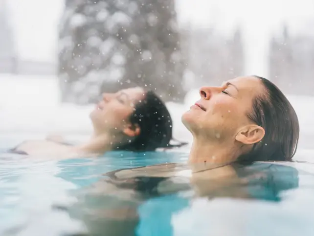 Two women lie back with their eyes closed in a warm, steaming pool and snow falls from the sky.