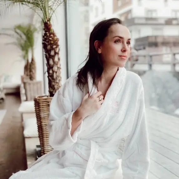 A woman in a bathrobe sits on a windowsill and looks through a window.