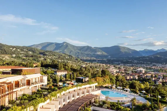 A view of the picturesque town of Salo on Lake Garda, surrounded by rolling hills and dense greenery. The architecture of the buildings on the shore conveys a classic Italian atmosphere, while the mountains in the background evoke a sense of adventure and closeness to nature. 