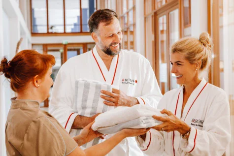 A couple in white bathrobes are handed towels by a member of staff.