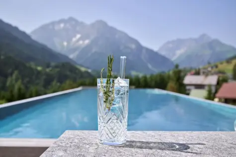An dem Beckenrand eines Pools steht ein Gin Tonic. Die Sonne scheint warm auf das Wasser und im Hintergrund sind Berge zu sehen. 