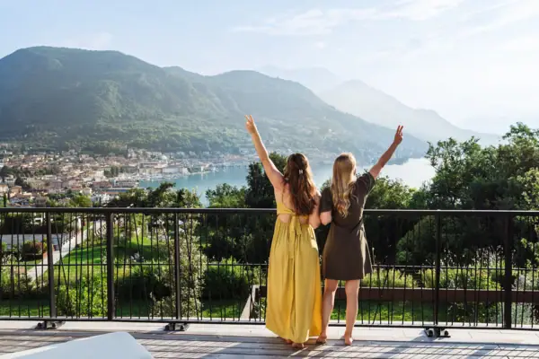 Zwei Frauen stehen auf einem Balkon mit Blick über den Gardasee und heben die Arme in die Luft.