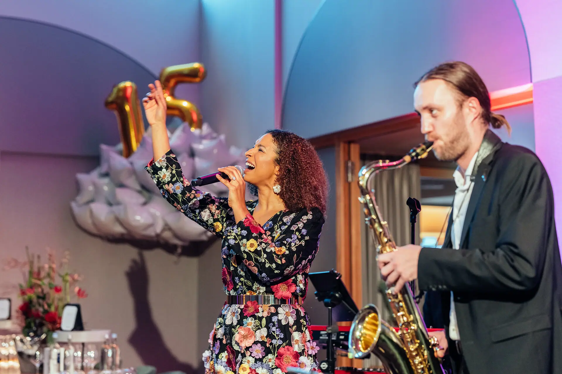 A woman is singing into a microphone whilst a man plays the saxophone.