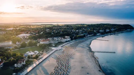 An aerial view of the coastal area of Travemünde. The water glows a warm blue, the beach stretches along the coast and many beach chairs are scattered along it, while the town lies quietly in the background among the greenery. 