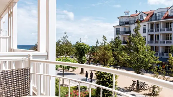 Balcony with white railing, overlooking a promenade with trees and a building in the background.