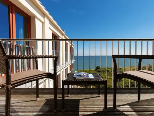 On a balcony there are two chairs and a small table with magazines on it from which you can look out over the sea. 