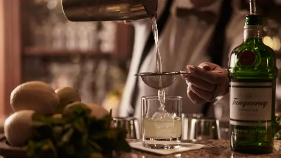 Person pours liquid from a cocktail shaker through a strainer into a glass.