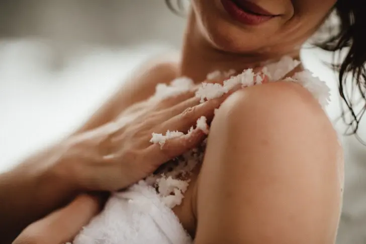 Close-up of a woman applying a salt scrub to her shoulder and arm. The grain of the scrub is clearly visible and emphasizes the care routine. Her relaxed smile indicates a wellness experience.