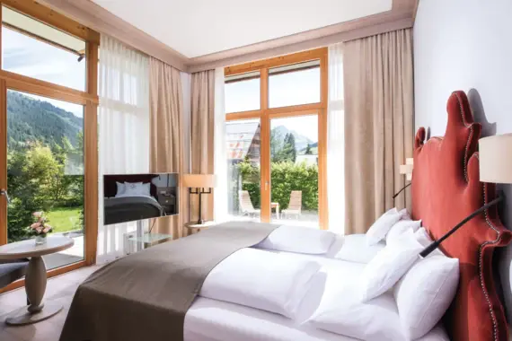 A bed in a room with a TV and large corner windows with wooden frames and a view of a green mountain backdrop.