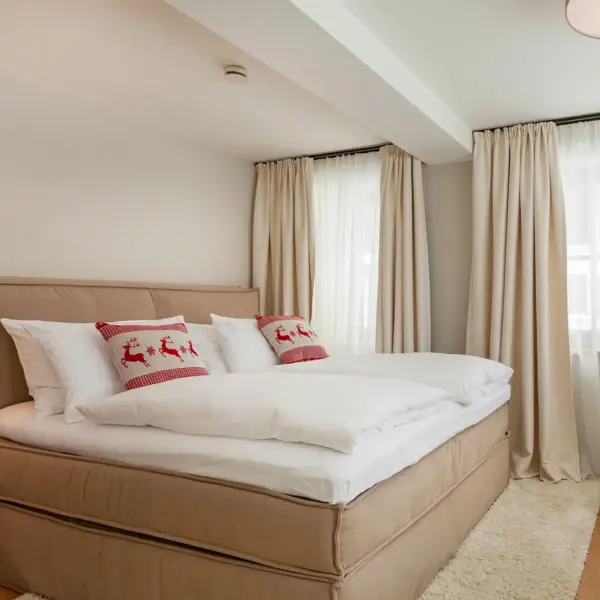 A large, cozy upholstered bed with white bed linen and two red decorative cushions with reindeer on them.