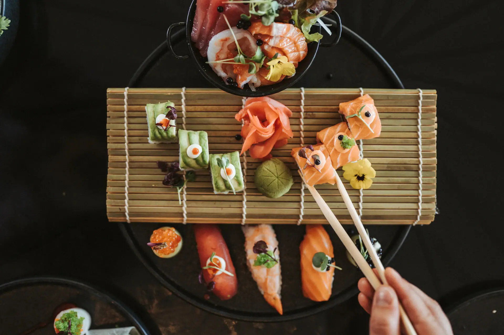 Person hält Essstäbchen zu einem Teller Sushi.