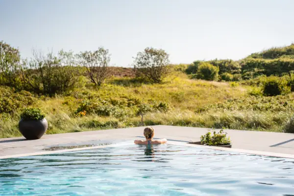 Eine Frau lehnt sich an den Beckenrand von einem Außenpool und schaut auf eine Dünenlandschaft.
