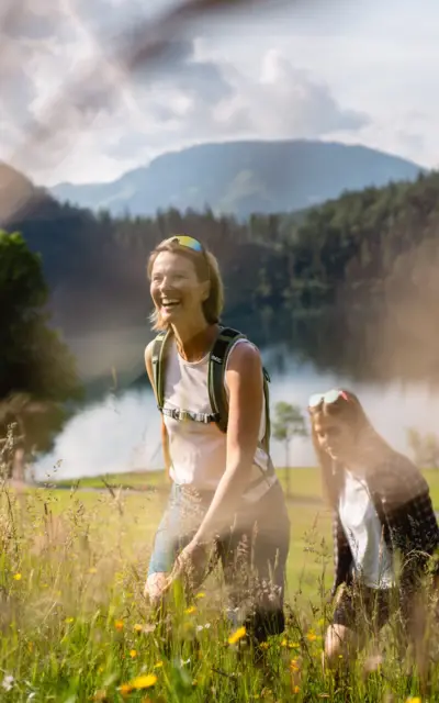 Two women are walking in a field, surrounded by grass and flowers against a mountain backdrop.