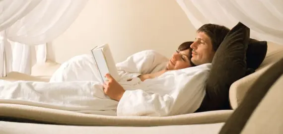 A man and a woman lie on a four-poster bed and read a book.