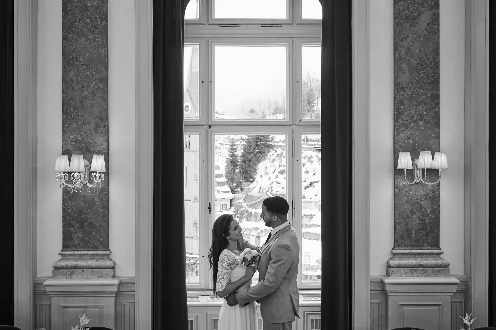 A man and woman standing in a room with a window, wearing formal clothing.