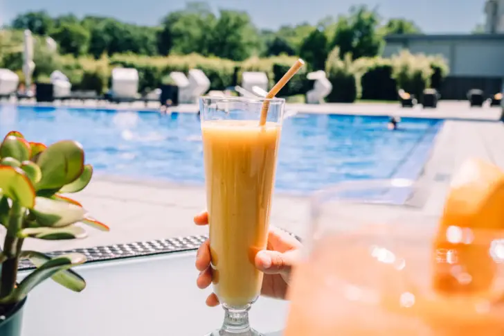A hand holds a yellow smoothie in a tall glass in front of an outdoor pool.
