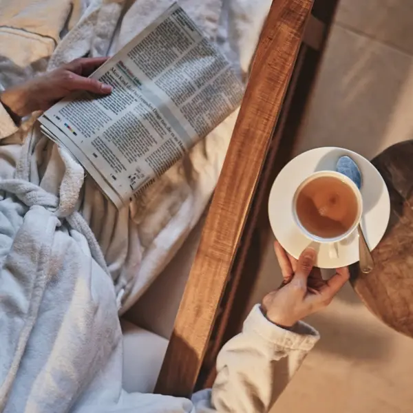 Person in a bathrobe holding a cup of tea and a newspaper in an interior room.