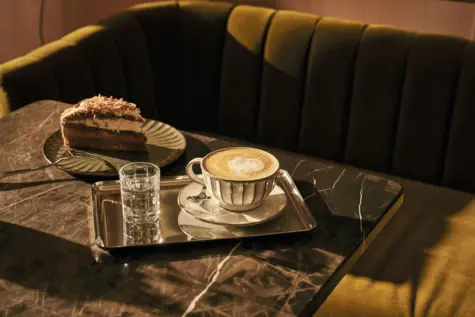 A laid table with a piece of cake and a small silver tray with a cup of cappuccino and a small glass of water.