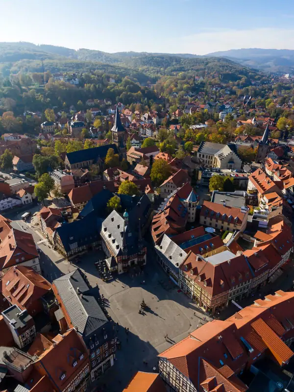 An aerial view of a town with houses and trees, surrounded by mountains and clouds in the sky.