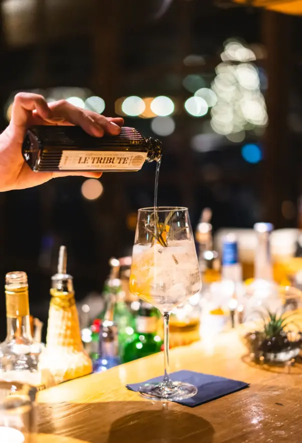 A hand pouring a drink from a small glass bottle into a glass draped on a wooden bar counter with bottles in the background. 