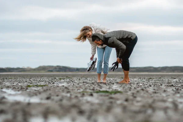 A man and a woman stand barefoot on the mudflats and look at the mud-covered ground. The sky is gray and the wind is blowing through their hair. 