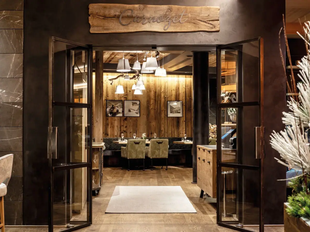 Ein gemütlicher Restaurantbereich mit vielen Holzdetails in Form von dem Boden sowie Holzwänden. Die Stühle und Bänke sind in natürlichen Grüntönen gehalten, an den Wänden hängen Bilder und weiße Lampenschirme, die von der Decke hängen, spenden Licht.