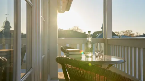 An inviting balcony with a round table and two wicker chairs, on which a bottle of water and glasses gleam in the sunlight. The warm rays of sunlight flood the scene and emphasize the relaxed atmosphere. The blue sky above the rooftops of the city can be seen in the background.