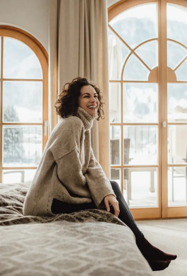 A woman in a cozy knitted sweater is sitting on abed smiling.Daylight enters the room through the large windows at the back and the mountains can be seen.