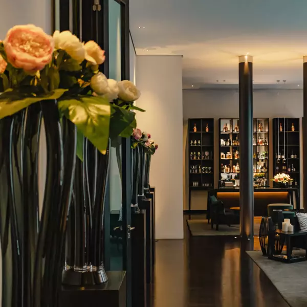 Several vases of dark smoked glass filled with white and orange peonies are lined up along the left edge. A bar can be seen in the background behind a round column. On the right in front of it is a seat.