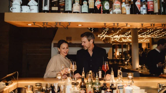 A couple is enjoying some white wine in a cozy looking place at the bar. 