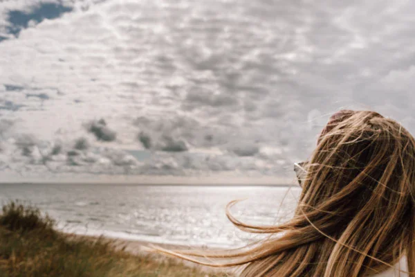 A woman looks out over the vastness of the sea in front of her while the sun shines warmly on the water and her hair blows in the wind. 