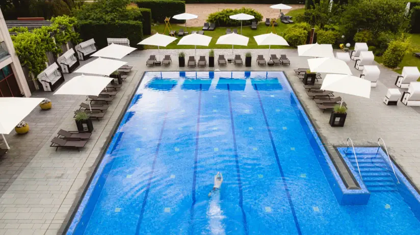 Aerial view of an outdoor pool in which a person is swimming. There are sunbeds and umbrellas around the pool 