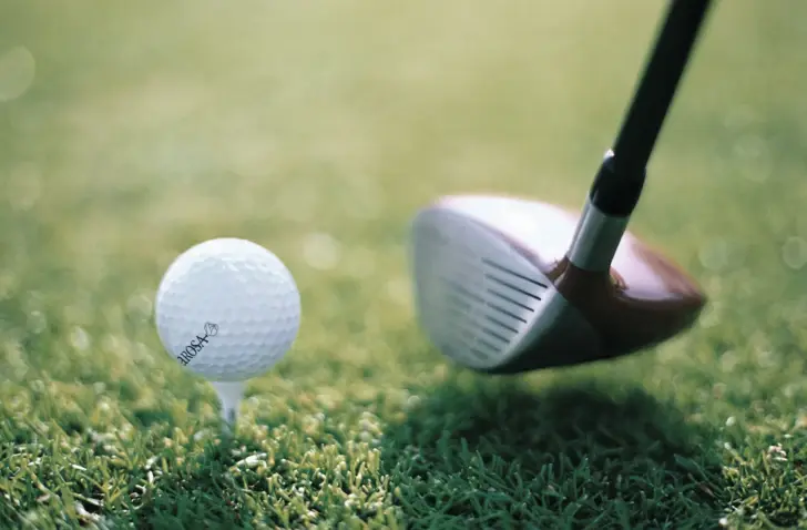A white golf ball is placed on a green gold square, which is aimed at with a golf club. 