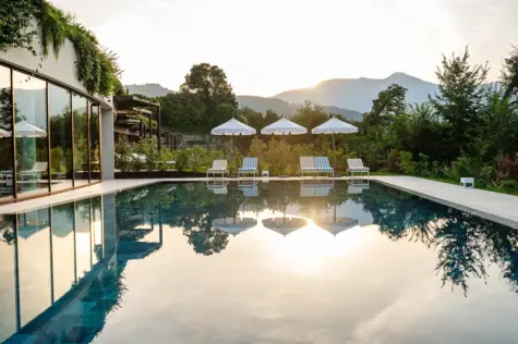 Outdoor swimming pool surrounded by chairs and parasols with trees and a clear sky with the setting sun in the background.