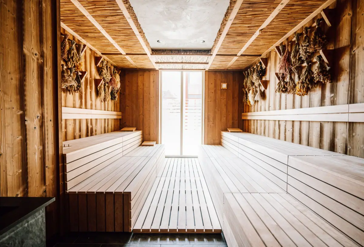 Ein Innenraum einer Sauna mit Holzbänken und einer Tür und getrockneten Kräutern, die Kopfüber an den Holzwänden hängen..