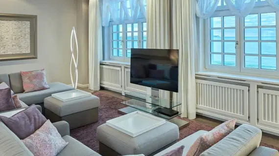 An elegant living room in a suite with a view of the sea. The furnishings consist of grey sofas and stools with color-coordinated cushions. In the center is a modern glass and white coffee table with a TV placed above it. A creative floor lamp adds a design touch, while the classic white curtains reveal traditional white window frames with a view of the Baltic Sea. An abstract piece of art hangs above one of the sofas, emphasizing the simple elegance of the room.