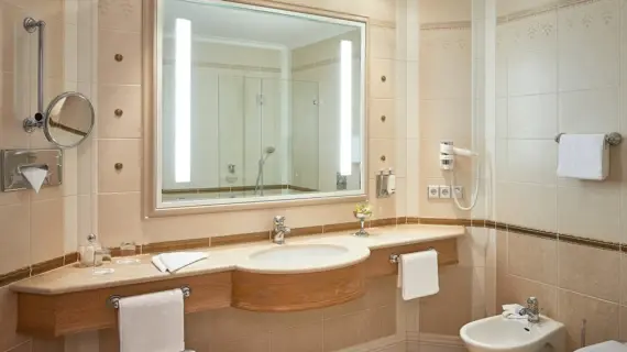 A bright, well-lit bathroom with beige tiles and matching borders. A large mirror above the washbasin reflects a glass shower screen. Below the mirror is a wooden washbasin with a marble top and a modern tap. There is a bidet next to the washbasin and a magnifying mirror and hairdryer on the wall. Fresh towels and a small vase with a flower create a homely atmosphere.