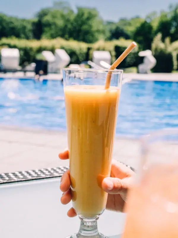 Eine Person hält ein Glas Orangensaft vor einem Außenpool im Hintergrund..