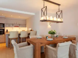 A kitchen and dining area in a vacation apartment. In the background you can see a modern kitchenette with three bar stools and in the foreground a large dining table with 5 armchairs.