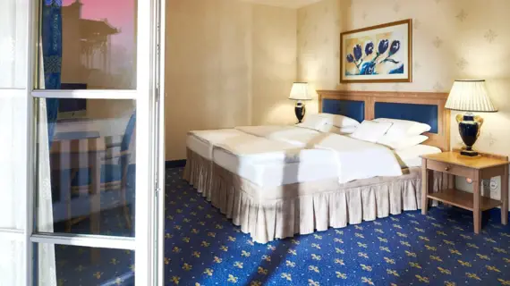 An elegantly designed double room in a hotel that radiates comfort with its warm colors and classic furnishings. The large bed with pristine white bed linen and a quilted headboard in soft blue tones is the centerpiece of the room. Above it hangs a colorful painting that is color-coordinated with the blue accents in the room. The bedside tables are fitted with stylish lamps whose soft light creates a relaxed atmosphere. The floor is covered with a blue carpet featuring small yellow floral motifs. An open French window with white bars suggests a connection to a balcony.