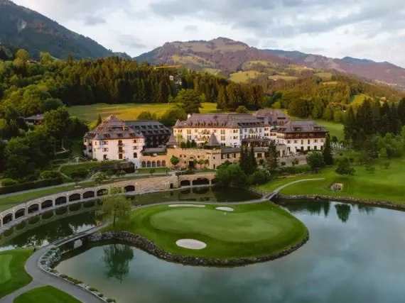 A golf course featuring a large building and a lake surrounded by trees and mountains.