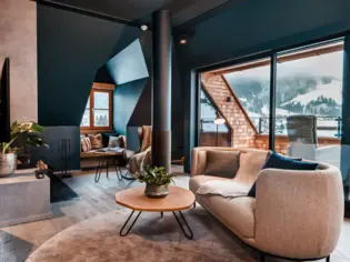 A bright living area with a gray sofa and a small round wooden table on a gray carpet. In the background you can see another seating area, a TV and a roof terrace.