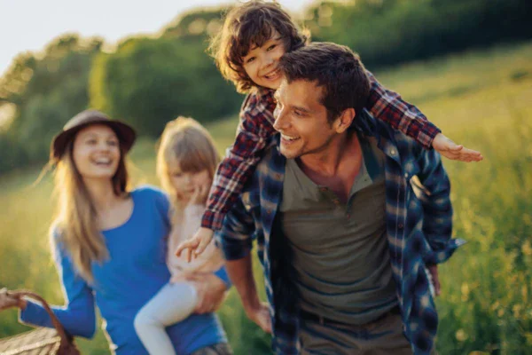A family walks happily along a country lane. The father is carrying his son on his back and the mother behind him is holding her little daughter in her arms. Green meadows and trees can be seen in the background. 