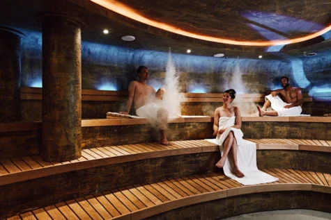 Three people are sitting in a sauna with wooden seats and stone features on the wall, ceiling and columns. Dimmed lights are shining and steam is coming from the ceiling. 