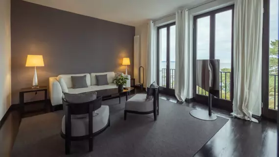 A living area with floor-to-ceiling windows and light-colored curtains. Small lamps bathe the room with sofa and armchairs as well as a TV in warm light.  