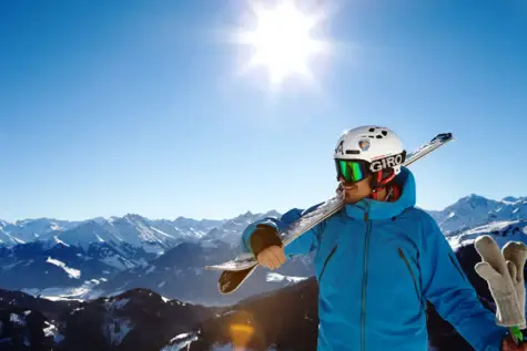 Man with ski helmet and goggles holding skis on his shoulder on the summit in front of a snowy mountain backdrop and blue sky with bright sun.