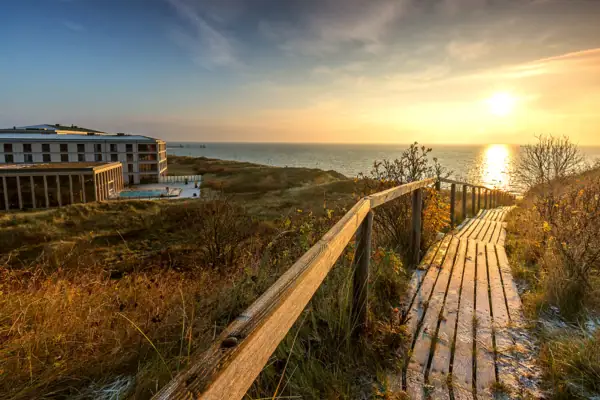 A wooden jetty leads to the beach, with a building and water in the background.