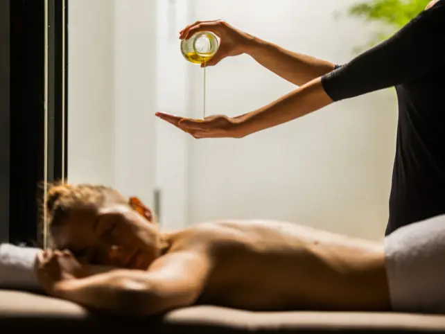 A woman lies relaxed on a massage table and above her the masseuse pours oil from a small false into her hand. 