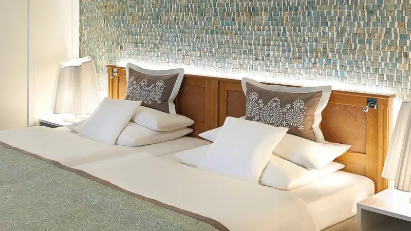 Rooms and Suites A neatly made bed with pillows and a duvet in a well-designed hotel room.
