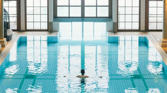 A woman is swimming in a pool that is surrounded by lid up columns and lounge seats. Big windows fill the SPA with daylight and a big door connects the pool with the outdoor area. 